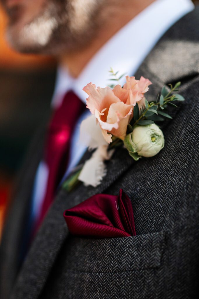 Close-up of a groom’s boutonniere featuring a peach flower, white ranunculus, and greenery.