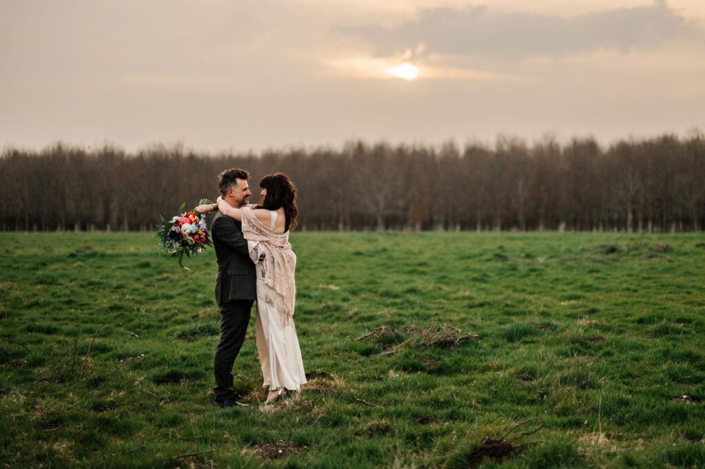 Bride and groom embracing in a grassy field at sunset with a colourful wedding bouquet.