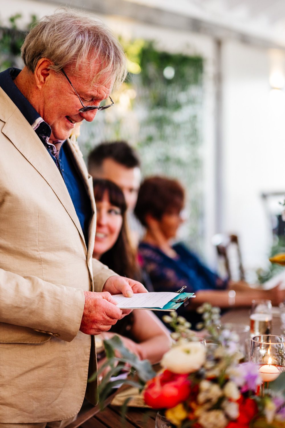 Wedding guest giving a speech at the reception with colourful floral arrangements on the table.